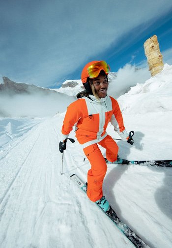 Lyžařský oblek v jasně oranžové s bílými akcenty. Tmavé rukavice. Lyže jsou černé se tyrkysovým spodkem. Sněhové pozadí s oblačnou oblohou.