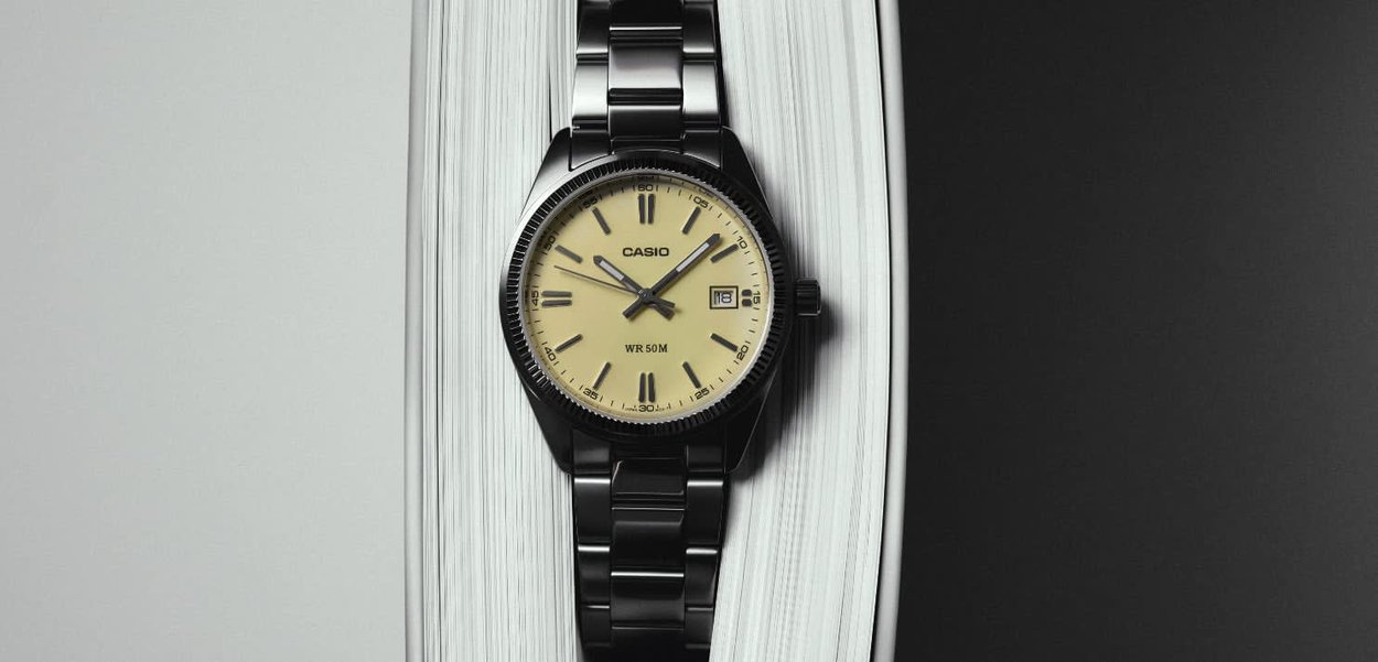 Reloj de pulsera Casio de metal negro con esfera beige, números romanos, visualización de la fecha y tres manecillas, descansando en el borde de un libro abierto.