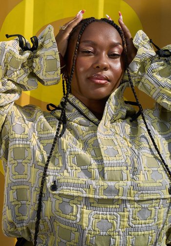 Gevoerde jas in lichtgeel en grijs met een geometrisch patroon. Voorzien van een hoge kraag en trekkoorden in de mouwen. Glad textuur, knoopsluiting.