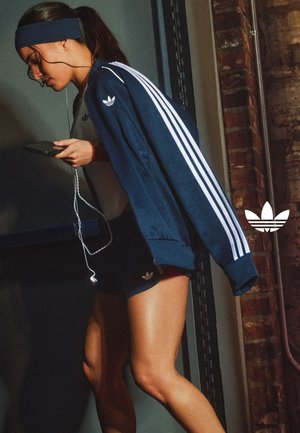 Kobieta w niebieskiej kurtce i spodenkach Adidas, trzymająca telefon ze słuchawkami, pochylona do przodu w pomieszczeniu blisko ceglanej ściany.
