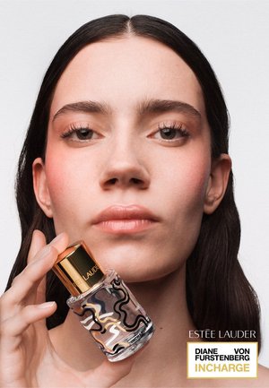 Frau mit langen, dunklen Haaren hält eine Estee Lauder Parfümflasche mit abstraktem schwarz-goldenem Design nahe an ihr Gesicht vor schlichtem Hintergrund.
