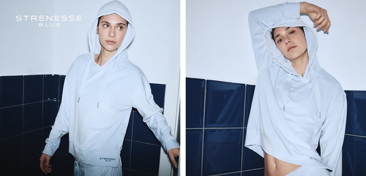 Frau trägt hellblauen Strenesse Blue Hoodie und posiert vor dunkler, blau gefliester Wand, zeigt entspannte und selbstbewusste Ausstrahlung.