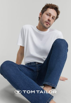 White cotton t-shirt and dark blue denim jeans, sitting posture against a plain background. Logo visible at the bottom.