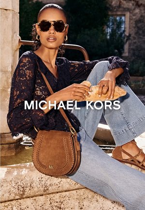 Donna con occhiali da sole e top in pizzo seduta su un muretto di pietra, che tiene in mano una baguette, con borsa a tracolla Michael Kors marrone e sandali.