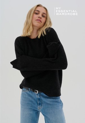 Pull en maille noire à coupe décontractée et manches longues, associé à un jean denim bleu clair avec une ceinture noire et boucles argentées.
