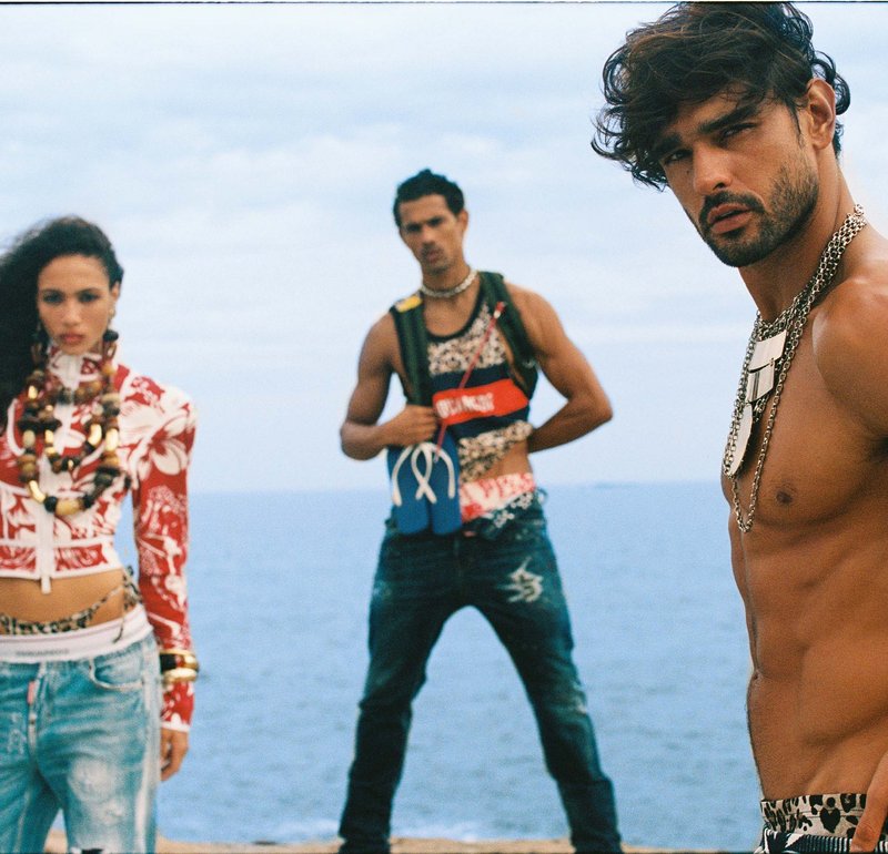 Tres jóvenes adultos con conjuntos de moda estampados están junto al mar, con un hombre sin camiseta delante y dos personas detrás de él.