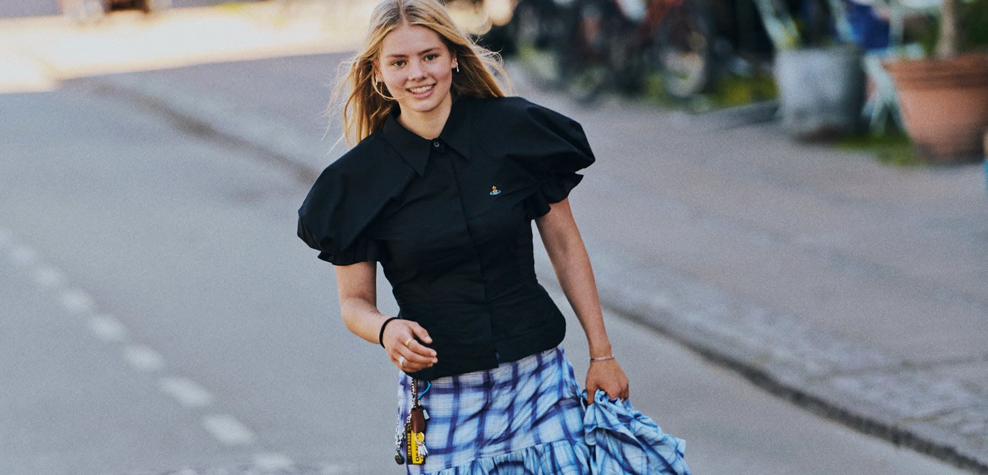 Ung kvinde smilende mens hun skateboarder på en gade iført en sort puffærmet top, en blåternet ruffle nederdel og sorte støvler.