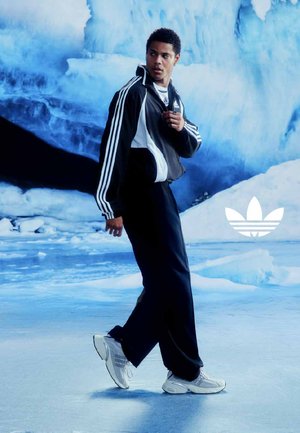 Man in zwart-wit Adidas trainingspak en witte sneakers die op een ijzige ondergrond loopt, met op de achtergrond een grote blauwe ijsformatie.