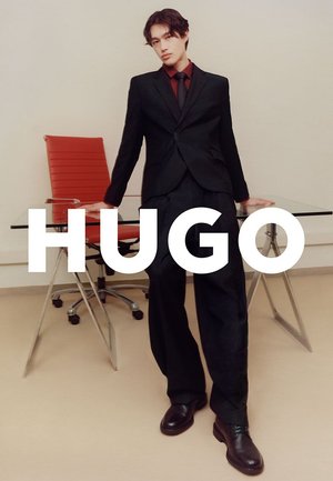 Schwarzer, maßgeschneiderter Anzug mit schmaler Passform, kombiniert mit einem dunkelroten Hemd und schwarzer Krawatte. Lederschuhe. Glasklarer Glastisch, roter ergonomischer Stuhl im Hintergrund.