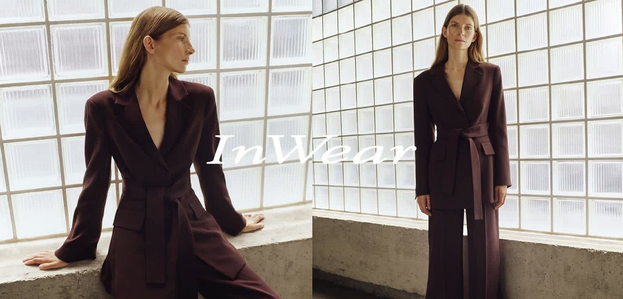 Frau mit langen braunen Haaren, die einen dunkel geschnittenen Anzug mit tailliertem Blazer trägt, steht und lehnt sich an eine Fensterbank aus Glasbausteinen.