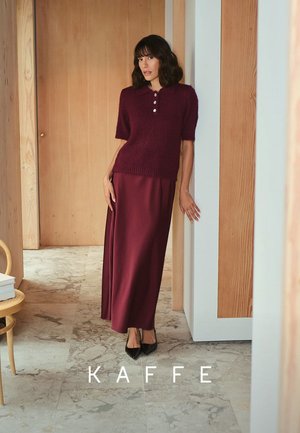 Burgundy knit short-sleeve polo with buttons, paired with a matching long satin skirt. Smooth texture, minimalist design, against a neutral backdrop.