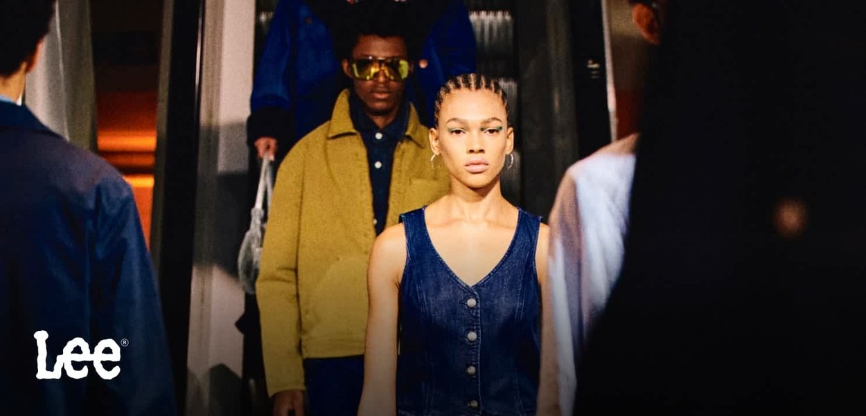 Robe salopette en denim bleu avec des boutons, portée par un mannequin. L'arrière-plan présente des personnes en vestes colorées et en accessoires. Marque Lee visible.