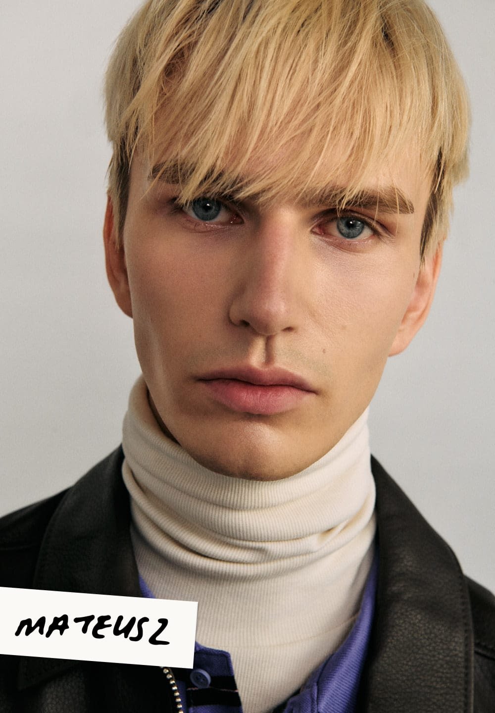 Portret osoby z blond włosami i niebieskimi oczami, ubranej w biały golf i czarną skórzaną kurtkę, na neutralnym tle.