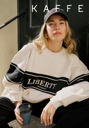 Frau mit schwarzer Kappe und weißem Liberty University Sweatshirt, hält eine Kaffeetasse und sitzt vor einer dunklen Tür mit dem Schriftzug "KAEFFE" darüber.