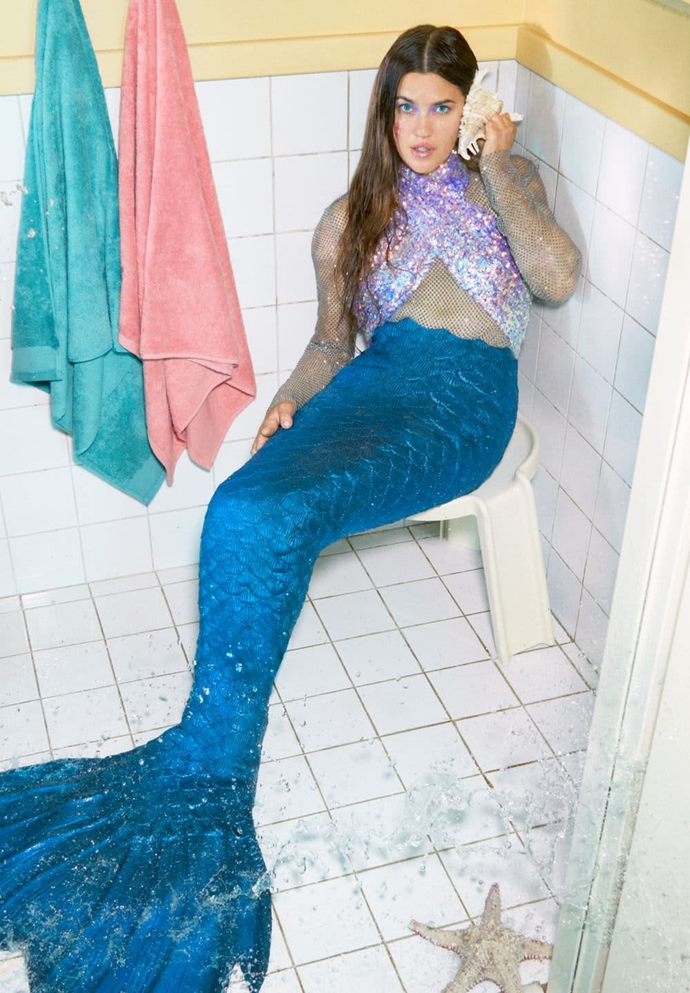 Mermaid costume with a sequined top and a textured blue tail, sitting on a white chair in a tiled bathroom with hanging towels.