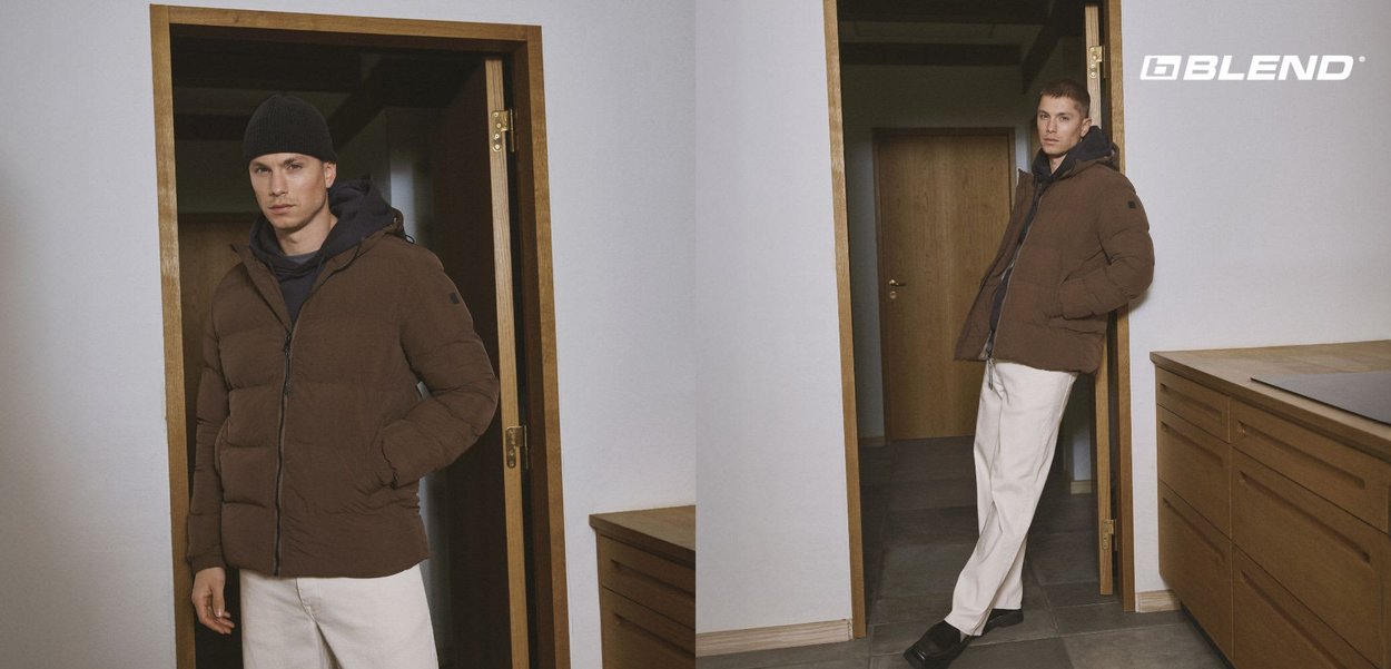 Giacca imbottita marrone con chiusura a zip e polsini elasticizzati, indossata sopra una felpa con cappuccio blu navy. Cappello lavorato a maglia nero completa il look. Sfondo interno piatto.