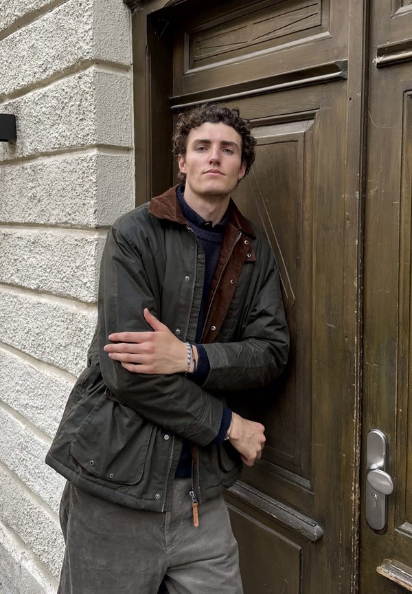 Donkergroene gewaxte jas met een bruine corduroy kraag, gedragen over een marineblauwe trui, gecombineerd met grijze corduroy broek. Houten deur op de achtergrond.