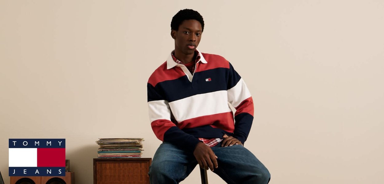 Polo à manches longues rayé rouge, marine et blanc avec col ; modèle portant un jean bleu assis sur un tabouret contre un mur beige.