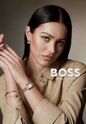 Femme aux cheveux lisses et foncés portant une chemise beige, une montre en or, des boucles d'oreilles créoles et des bracelets, posant avec les mains croisées près du visage.