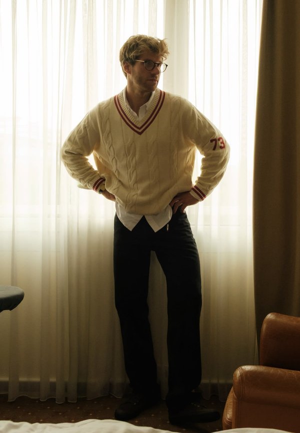 Suéter de punto trenzado color crema con acentos rojos, superpuesto a una camisa blanca con cuello, combinado con pantalones oscuros. De pie junto a cortinas translúcidas.