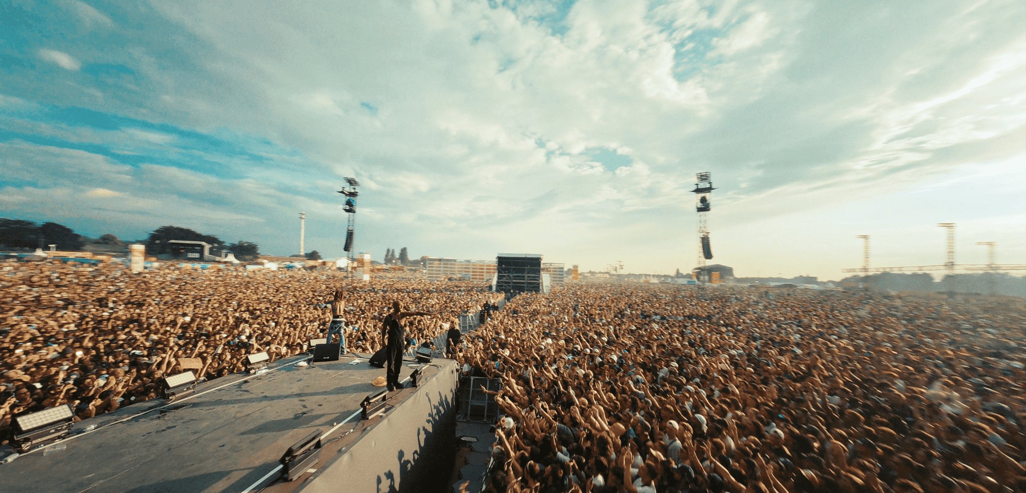Menigte op een muziekfestival onder een blauwe lucht, met een groot podium en apparatuur op de voorgrond, verlicht door de lichten van het podium.