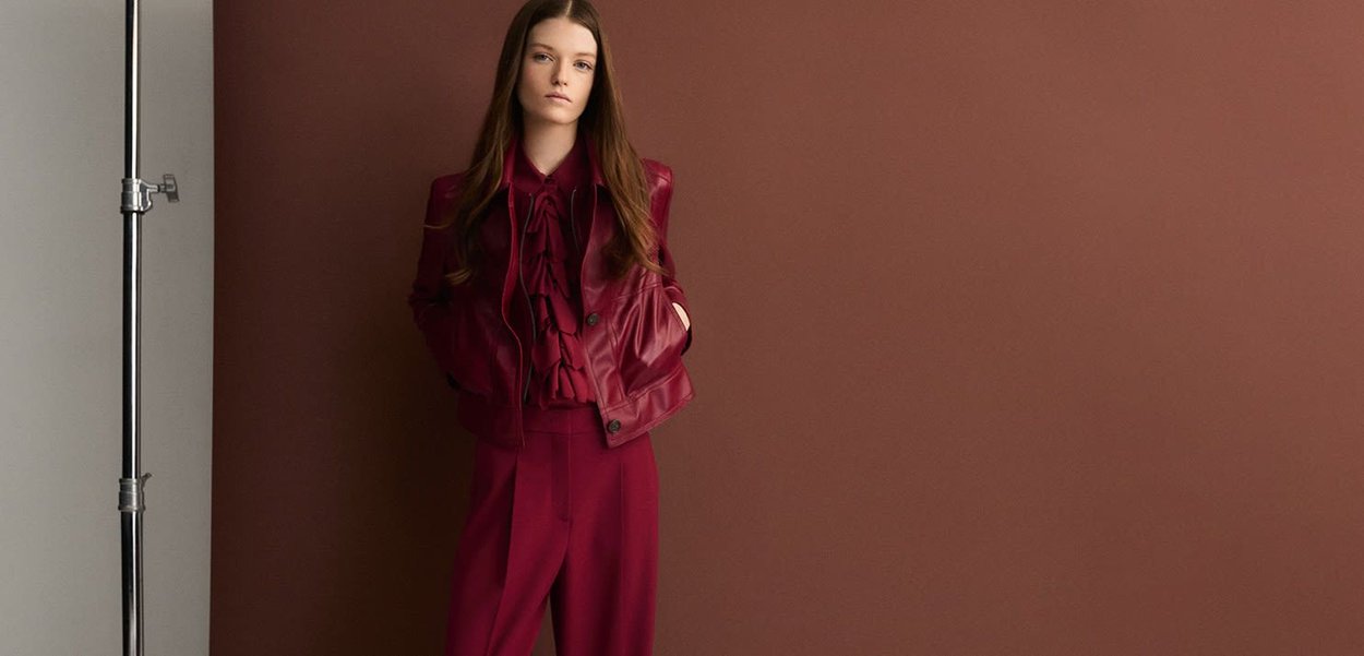 Burgundy leather jacket with ruffled blouse and matching wide-leg trousers. Smooth texture, relaxed fit, set against a dark brown background.