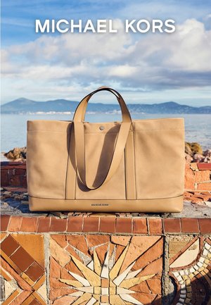 Skinnveske i tan fra Michael Kors, hvilende på en murvegg kledd med mosaikkfliser, med sjø og fjell i bakgrunnen.