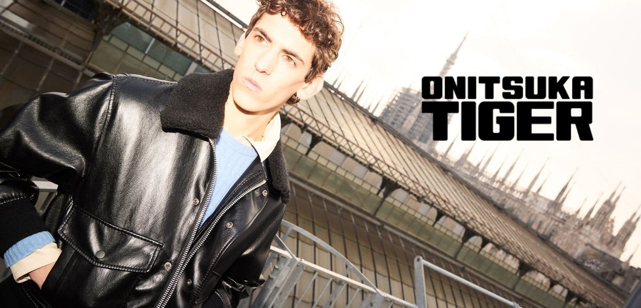 Schwarze Lederjacke mit Fellkragen, ausgestattet mit Reißverschluss und Klappentaschen, über einem hellblauen Pullover getragen. Urbaner Hintergrund.