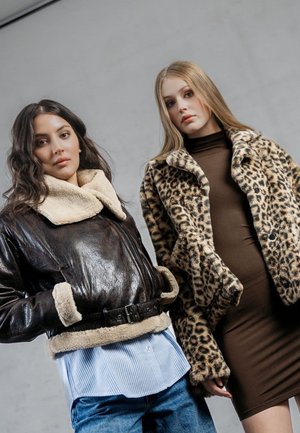 Brązowa skórzana kurtka z kołnierzem z owczej wełny i paskiem; futrzany płaszcz w leopardzie; obie modelki noszą dopasowane bluzki i luźne dżinsy.