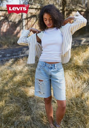 Vrouw buiten die een wit hemdje, gestreepte jas en gescheurde knielange spijkershorts met het Levi's-logo in de hoek draagt.