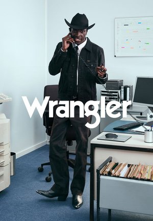 Czarna dżinsowa kurtka na białej koszuli, czarne spodnie i czarny kapelusz kowbojski. Stoi w biurze z telefonem, otoczony meblami biurowymi.