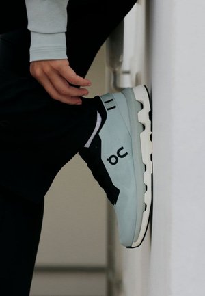 Person, die hellblau-schwarze Laufschuhe mit dem "On"-Markenlogo trägt und ein Bein an einer weißen Wand dehnt.