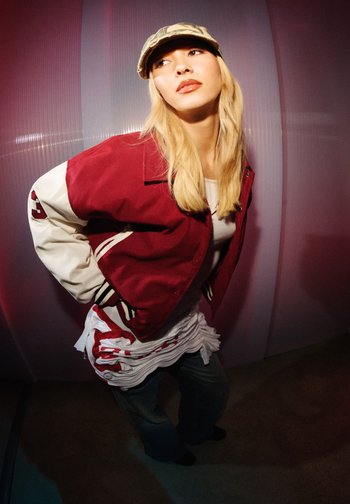 Joven con cabello largo y rubio está de pie frente a una pared morada, vistiendo una chaqueta roja y blanca, camiseta clara, jeans y una gorra de camuflaje.