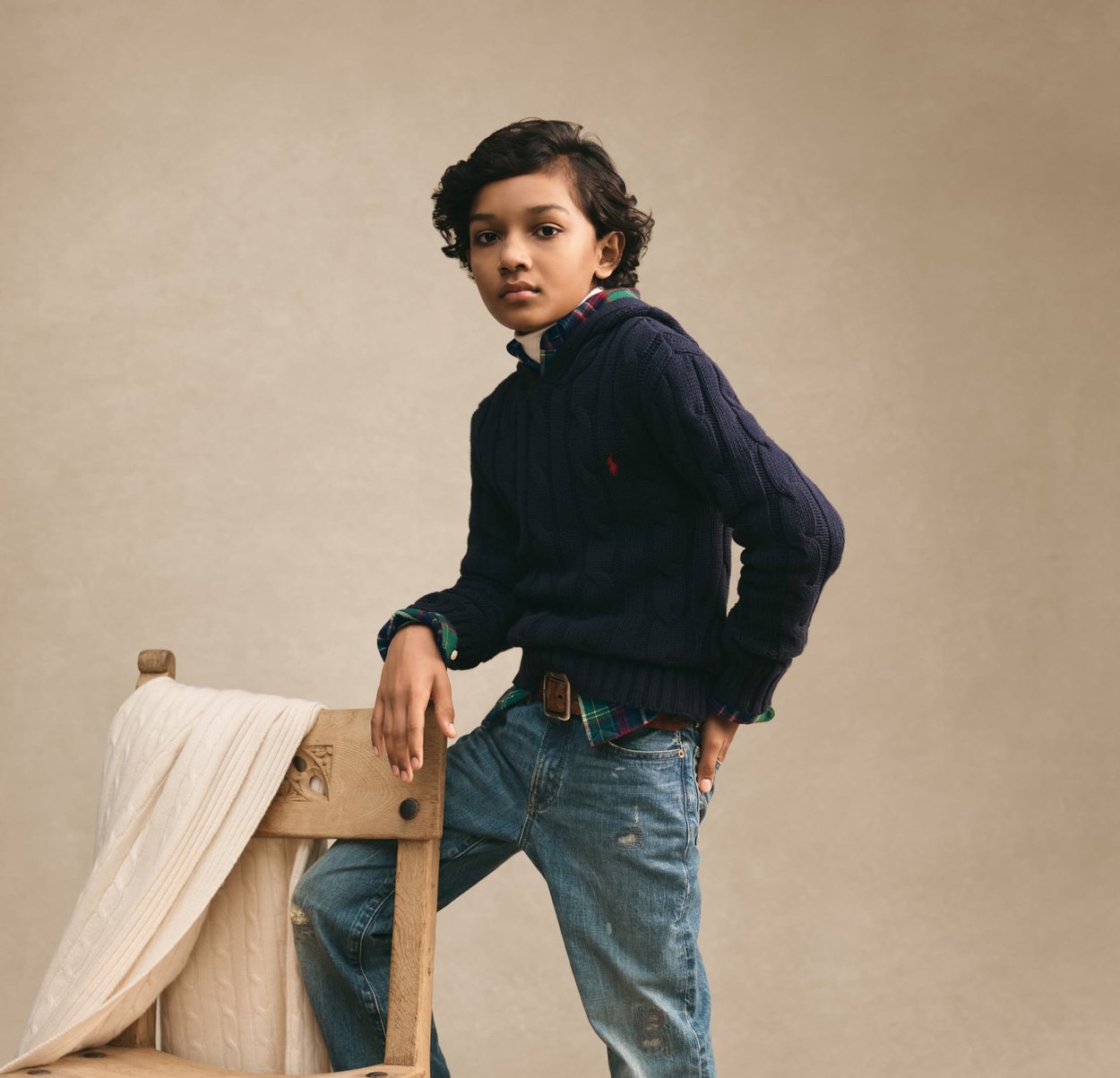 Enfant aux cheveux bouclés foncés portant un pull marine et un jean, appuyé sur une chaise en bois recouverte d'une couverture en tricot crème, sur un fond neutre.