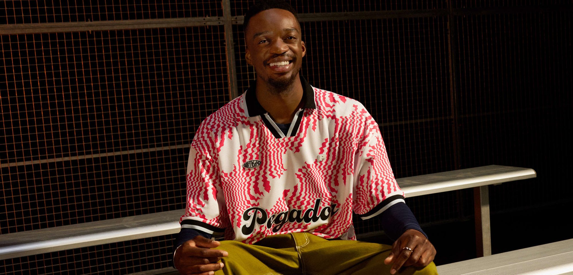 Buntes Kurzarmtrikot mit roten abstrakten Mustern, schwarzem Kragen, kombiniert mit olivgrünen Hosen und schwarz-gelben Sneakern. Silberne Bank.