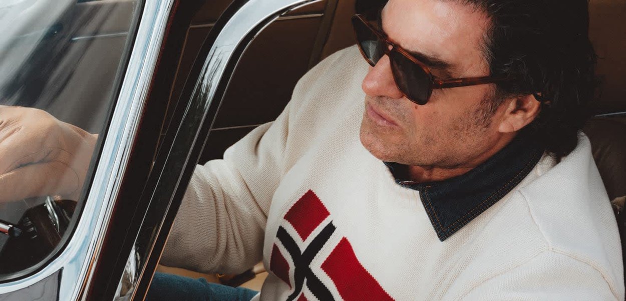 Uomo con occhiali da sole e maglione bianco con motivo rosso e nero, seduto in macchina e con le mani sul volante.