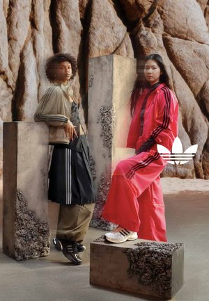 Zwei Frauen in sportlichen Adidas-Outfits posieren neben strukturierten Betonblöcken vor einem felsigen Hintergrund, eine in Schwarz und Beige, die andere in lebhaftem Rot.