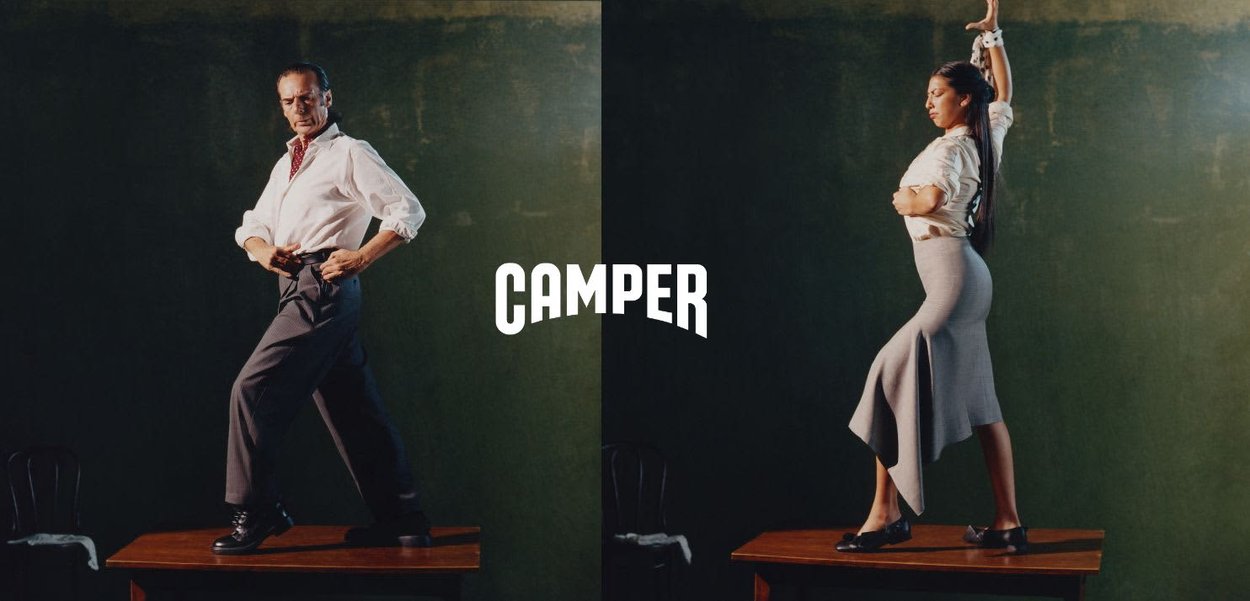 Mann und Frau stehen selbstbewusst auf Holztischen, tragen weiße Oberteile und dunkle Hosen, vor grünem Hintergrund mit zentriertem "Camper"-Logo.