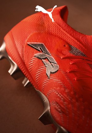 Scarpetta da calcio rossa brillante con materiale in mesh, marchio argento e superficie testurizzata. Presenta un design low-top e tacchetti in metallo per la trazione.