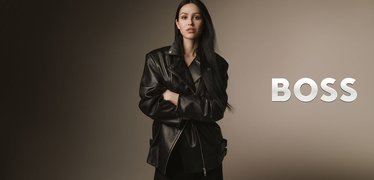 Czarna skórzana kurtka o kroju dwurzędowym, z dużymi klapami i ozdobami na zamek, noszona na całościowo czarnym stroju; tło neutralne.