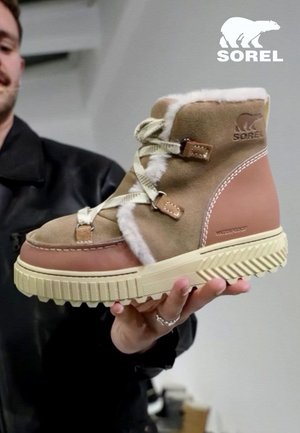Homme tenant une botte d'hiver Sorel beige et marron avec doublure en fourrure, semelle épaisse en caoutchouc et fermeture à lacets sur un fond uni.