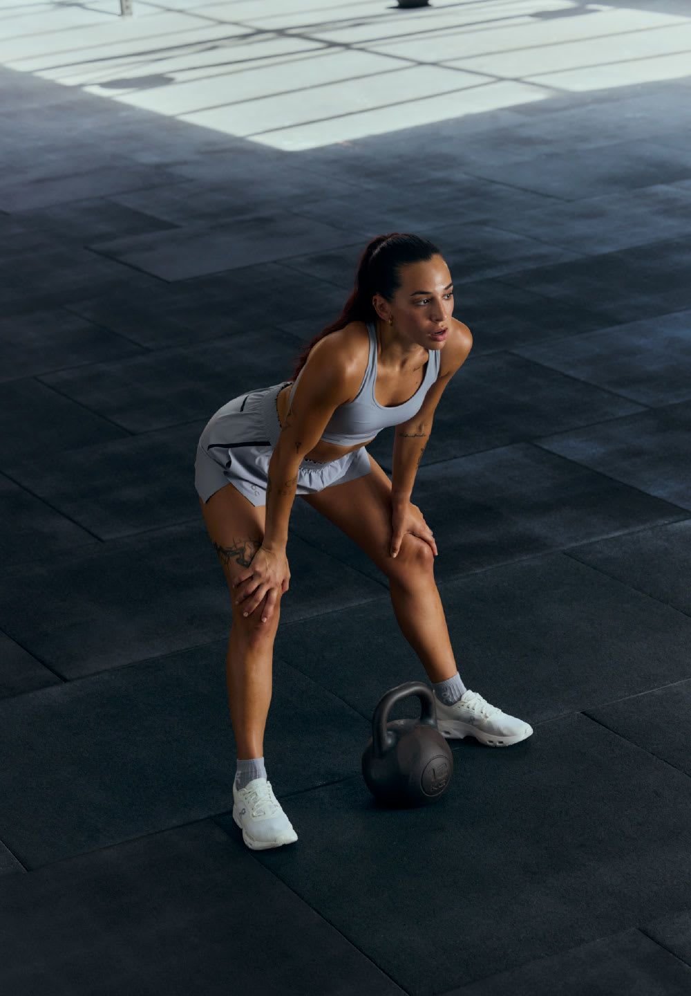 Sportliche Frau in grauer Sportbekleidung, die auf dem schwarzen Gummiboden des Fitnessstudios hockt, mit einer Kettlebell vor sich; ihre Schuhe sind weiß mit grauen Akzenten.