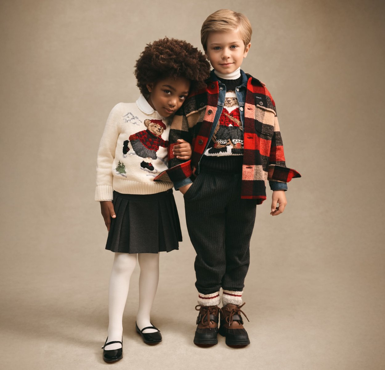 Zwei Kinder stehen. Ein Mädchen mit lockigem Haar, trägt einen weißen Pullover mit Bärenmotiv und einen Rock, ein Junge in geschichteter Kleidung mit kariertem Mantel und Bärenpullover.