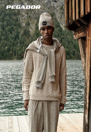 Beiger Strickpullover mit Kabelmuster und Rippstruktur, kombiniert mit passenden weiten Hosen und einer Mütze. Mit einem Logo akzentuiert. Natürliches Außenambiente.