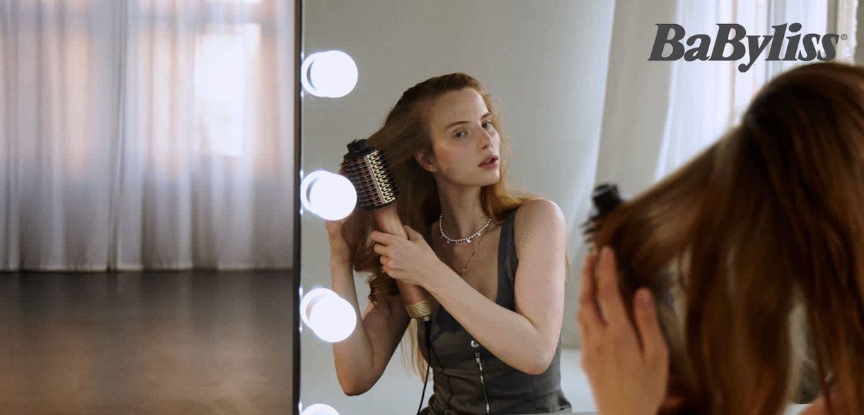 BaByliss hair styling brush with a metallic gold handle, black bristles, and a round shape. Clear reflections in a well-lit mirror setting.