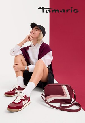 Baskets de sport rouge et beige avec des lacets blancs, accompagnées d'un sac à bandoulière bordeaux assorti. En arrière-plan, des blocs de couleur beige et rouge doux.
