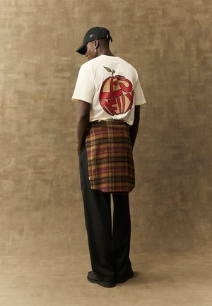 Hvid t-shirt med grafisk rødt æble og tekst på ryggen; brun ternet nederdel over sorte bukser; sort kasket og sko. Ensfarvet baggrund.