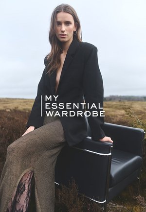 Schwarzer, maßgeschneiderter Blazer mit Revers und Kerbkragen, kombiniert mit einem strukturierten braunen Rock mit Spitzenakzenten, vor einer natürlichen Landschaft.