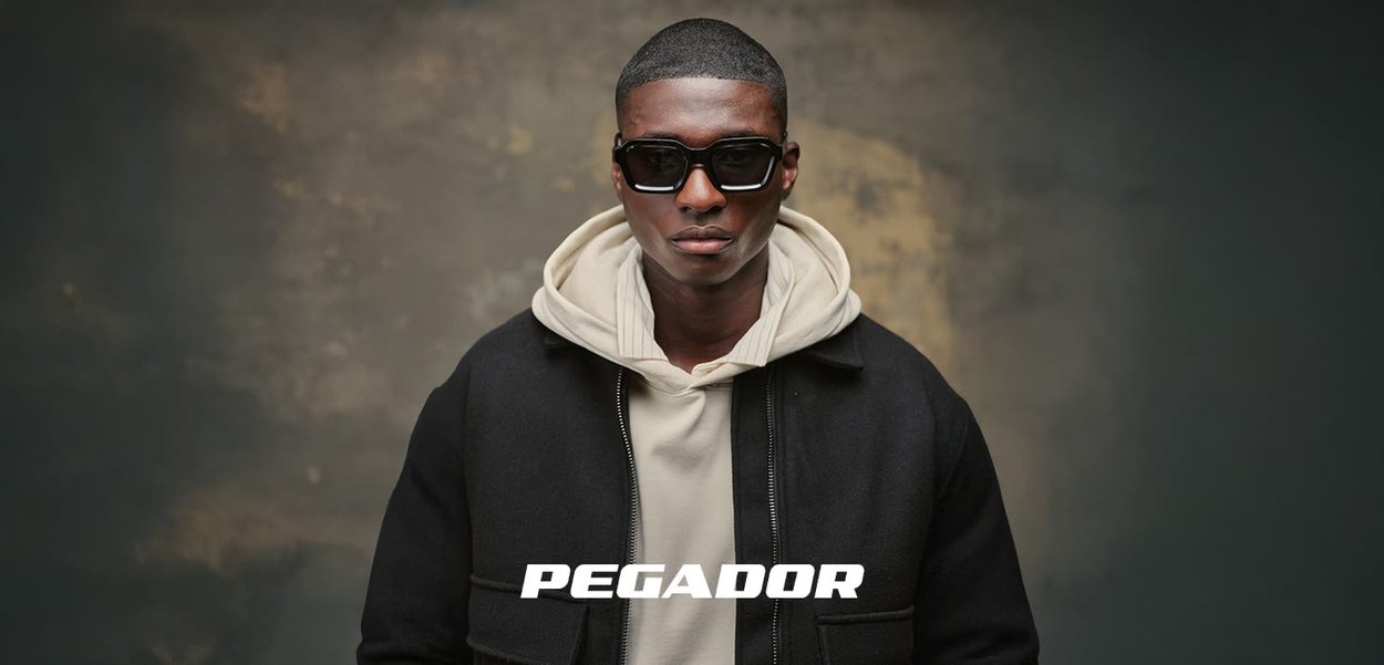 Junger Mann mit dunkler Sonnenbrille, cremefarbenem Hoodie und schwarzer Jacke steht vor einem strukturierten grauen Hintergrund mit dem Schriftzug "PEGADOR" darunter.