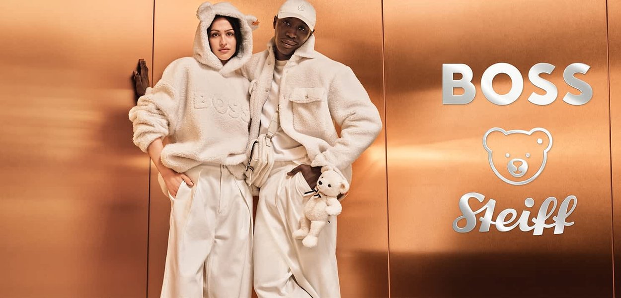 Zwei Modelle tragen cremefarbene, plüschige Outfits: einen Kapuzenpullover und eine Jacke. Einer hält einen kleinen Teddybären. Mattierte Metallhintergrund.